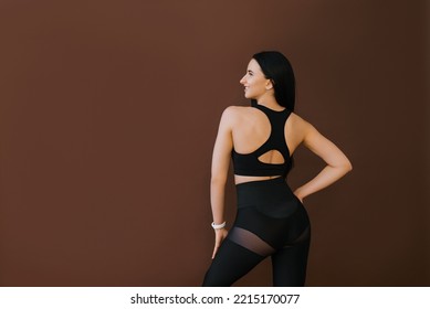 Rear View From Behind A Beautiful Curvy Sexy Young Brunette Fitness Trainer Posing Looking Up, Isolated On A Brown Background, Studio Portrait