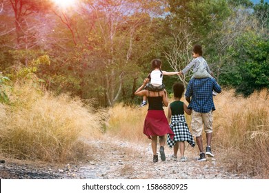 Rear View Of Big Mixed Race Family, Father And Mother Carrying Our Daughter Walking Along Yellow Field To Campsite. Travel Vacation Concept.