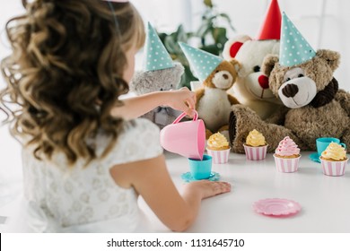 Rear View Of Birthday Kid Having Tea Party With Teddy Bears In Cones At Table With Cupcakes 