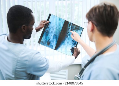 Rear View Of Black Doctor Holding X-ray Image Of Spine Against Window Light And Discussing It With Colleague