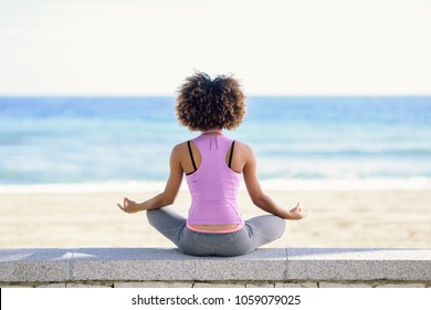 Rear view of black woman, afro hairstyle, doing yoga in the beach. Young Female wearing sport clothes in lotus figure with defocused background. - Powered by Shutterstock