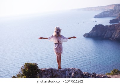 rear view of blond hair woman standing on the edge of a cliff and enjoing calm and silence, wearing in boho style closeses against the sea or ocean - Powered by Shutterstock