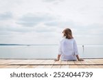 Rear view of a blond haired middle-aged woman sitting on the pier by the lake. Full length shot. Confident female wearing white shirt and blue jeans.