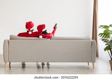Rear view of bonding black lovers in red Santa hats sitting on couch at home, watching TV, unrecognizable lady aiming remote controller towards copy space on white wall, panorama, full length