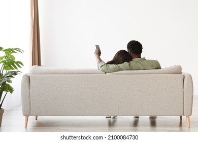 Rear view of bonding black lovers sitting on couch at home, watching TV, unrecognizable african american man aiming remote controller towards copy space on white wall, panorama, full length