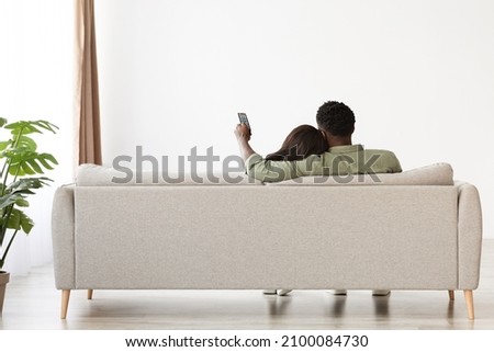 Rear view of bonding black lovers sitting on couch at home, watching TV, unrecognizable african american man aiming remote controller towards copy space on white wall, panorama, full length