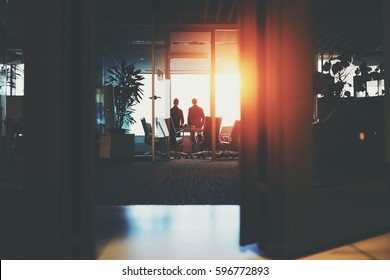 Rear View From Bottom Through Half-closed Doors Of Silhouettes Of Two Young Businessmen Near Big Window In Dark Office Interior After Meeting Having Rest, Discussing Something And Looking Outside