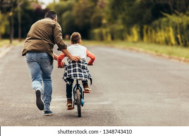 Rear view of a boy riding a bicycle while his father runs along holding the kid. Father teaching his son to ride a bicycle. - Shutterstock ID 1348743410