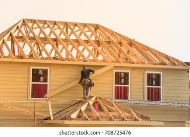 Rear View Of Builder At Eco Wooden House Construction Site Near Completed Suburban Home In Humble, Texas, US During Sunset. Carpenter On Scaffolding Installing Board Of Stick Built. Labor Day Concept