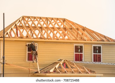 Rear View Of Builder At Eco Wooden House Construction Site Near Completed Suburban Home In Humble, Texas, US During Sunset. Carpenter At Work. Construction Worker Using Electric Drill On Building Site