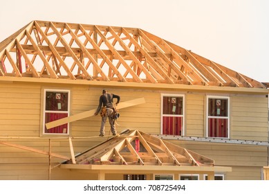 Rear View Of Builder At Eco Wooden House Construction Site Near Completed Suburban Home In Humble, Texas, US During Sunset. Carpenter On Scaffolding Installing Board Of Stick Built. Labor Day Concept