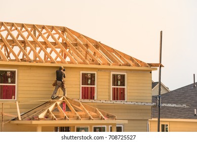 Rear View Of Builder At Eco Wooden House Construction Site Near Completed Suburban Home In Humble, Texas, US During Sunset. Carpenter At Work. Construction Worker Using Electric Drill On Building Site