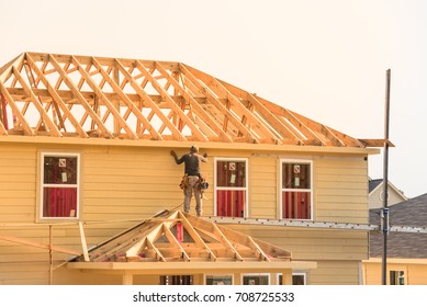 Rear View Of Builder At Eco Wooden House Construction Site Near Completed Suburban Home In Humble, Texas, US During Sunset. Carpenter On Scaffolding Installing Board Of Stick Built. Labor Day Concept