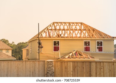 Rear View Of Builder At Eco Wooden House Construction Site Near Completed Suburban Home In Humble, Texas, US During Sunset. Carpenter On Scaffolding Installing Board Of Stick Built. Labor Day Concept