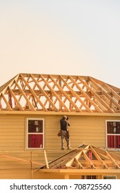 Rear View Of Builder At Eco Wooden House Construction Site Near Completed Suburban Home In Humble, Texas, US During Sunset. Carpenter At Work. Construction Worker Using Electric Drill On Building Site