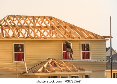 Rear View Of Builder At Eco Wooden House Construction Site Near Completed Suburban Home In Humble, Texas, US During Sunset. Carpenter At Work. Construction Worker Using Electric Drill On Building Site