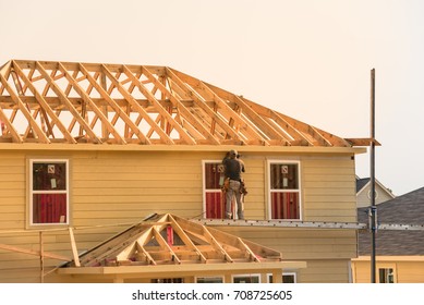 Rear View Of Builder At Eco Wooden House Construction Site Near Completed Suburban Home In Humble, Texas, US During Sunset. Carpenter At Work. Construction Worker Using Electric Drill On Building Site