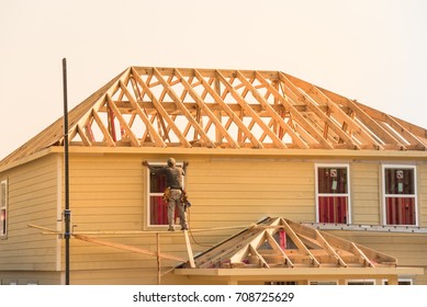 Rear View Of Builder At Eco Wooden House Construction Site Near Completed Suburban Home In Humble, Texas, US During Sunset. Carpenter On Scaffolding Installing Board Of Stick Built. Labor Day Concept