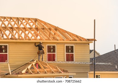 Rear View Of Builder At Eco Wooden House Construction Site Near Completed Suburban Home In Humble, Texas, US During Sunset. Carpenter At Work. Construction Worker Using Electric Drill On Building Site