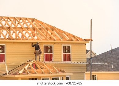 Rear View Of Builder At Eco Wooden House Construction Site Near Completed Suburban Home In Humble, Texas, US During Sunset. Carpenter On Scaffolding Installing Board Of Stick Built. Labor Day Concept