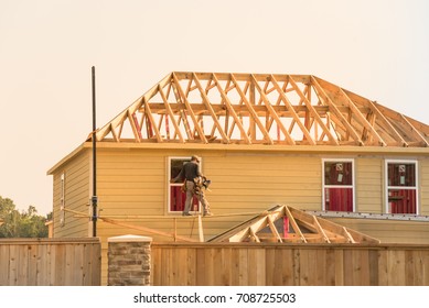 Rear View Of Builder At Stick Built Wooden House Construction Site Near Completed Suburban Home In Humble, Texas, US During Sunset. Framing Contractor Wears Tool Belt On Scaffolding. Labor Day Concept