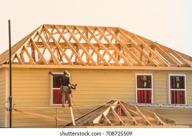 Rear View Of Builder At Stick Built Wooden House Construction Site Near Completed Suburban Home In Humble, Texas, US During Sunset. Framing Contractor Wears Tool Belt On Scaffolding. Labor Day Concept