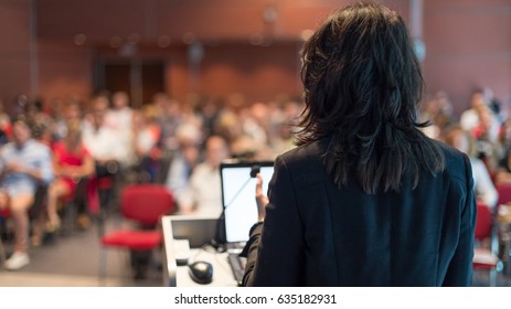 Rear view of business woman at lectern lecturing at Conference. Audience at the lecture hall.