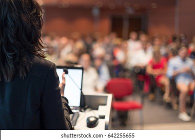 Rear view of business woman at lectern lecturing at Conference. Audience at the lecture hall.