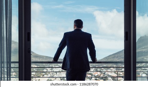 Rear View Of Businessman Standing By Hotel Room Balcony Looking At The City View.  Man In Business Suit Looking Outside From Hotel Room.