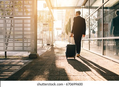 Rear View Of Businessman Walking With Bag Outside Airport. Young Business Traveler Pulling Suitcase On City Street.