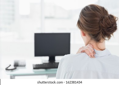 Rear view of a businesswoman with neck pain in front of computer in a bright office