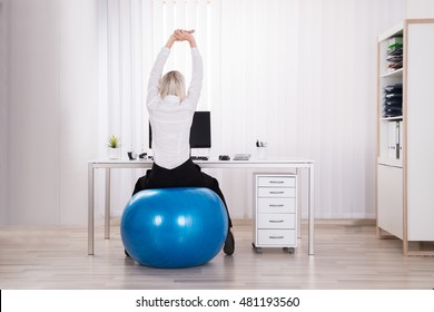 Rear View Of Businesswoman Sitting On Fitness Ball Stretching Her Arms In Office - Powered by Shutterstock