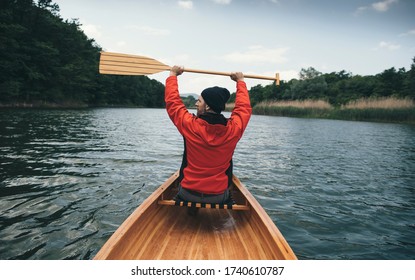 Rear view of canoeist in red jacket holding oars high. Man paddling canoe on cloudy day