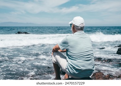 Rear view of carefree senior relaxed man barefoot wearing  cap sitting on a rocky beach admiring the sea waves crashing on the beach. Travel vacation leisure activity concept - Powered by Shutterstock