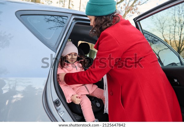 Rear view\
of a caring mom in bright red warm coat fastening her adorable\
little daughter\'s seat belt. Safe movement of children in the car,\
auto security and safety travel by car\
concept.