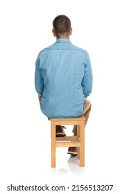 Rear View Of Casual Man In Denim Shirt Sitting On Wooden Chair In Front Of White Background In Studio