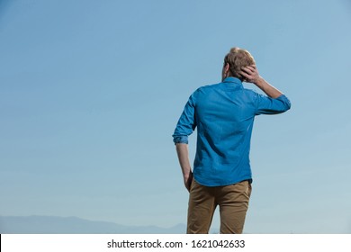 Rear View Of A Casual Man Standing With One Hand In Pocket And Scratching His Head While Pondering An Important Decision Outside On Blue Sky