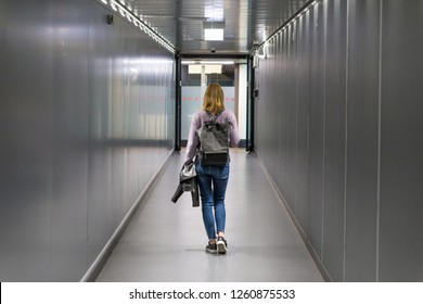 Rear View Of A Casual Woman Traveling, Wearing Small Travel Backpack, Piece Of Plane Hand Luggage, Walking Narrow Airport Corridor Leading To Baggage Pickup After Airplane Landing.