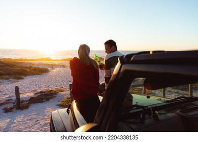 Rear view of a Caucasian couple outside their open top car, with sunset on the beach in the background, making a toast - Powered by Shutterstock