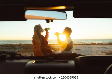 Rear view of a Caucasian couple outside their open top car, with sunset on the beach in the background, drinking beer  - Powered by Shutterstock