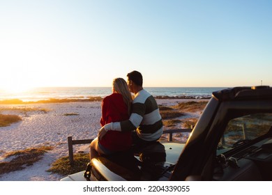 Rear view of a Caucasian couple outside their open top car, with sunset on the beach in the background, embracing and looking away - Powered by Shutterstock