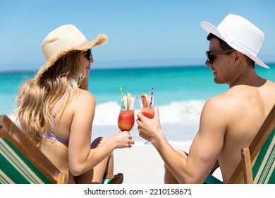 Rear view of a Caucasian couple sitting on deckchairs on the beach with blue sky and sea in the background, smiling, looking at each other and making a toast  - Powered by Shutterstock