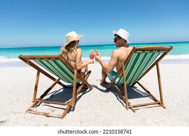 Rear view of a Caucasian couple sitting on deckchairs on the beach with blue sky and sea in the background, smiling, looking at each other and making a toast  - Powered by Shutterstock