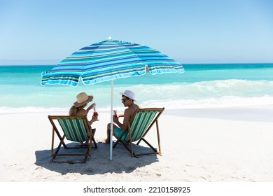 Rear view of a Caucasian couple sitting on deckchairs under the umbrella, on the beach with blue sky and sea in the background, smiling, looking at each other and making a toast  - Powered by Shutterstock