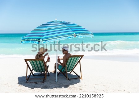 Rear view of a Caucasian couple sitting on deckchairs under the umbrella, on the beach with blue sky and sea in the background, smiling, looking at each other and making a toast 