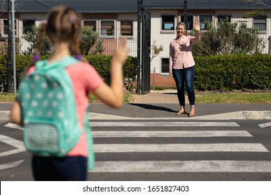 Rear view of a Caucasian schoolgirl wearing a backpack waving to her mother from the other side of a pedestrian crossing on her way to elementary school on a sunny day