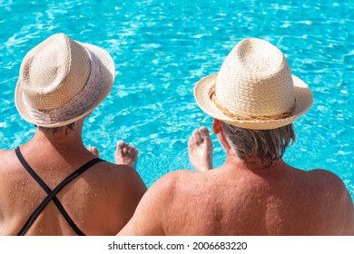 Rear View Of Caucasian Senior Couple Sitting On The Edge Of The Pool With Their Feet In The Water. Two Happy Retirees Enjoy Their Summer Holidays Under The Sun