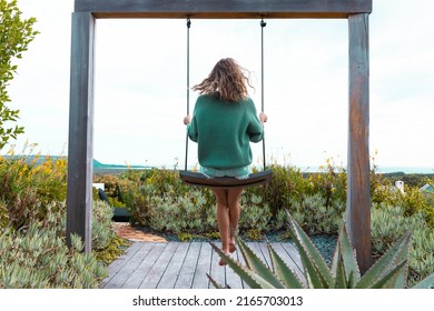 Rear view of caucasian woman sitting on swing amidst plants against clear sky, copy space. nature, unaltered, lifestyle, enjoyment and holiday concept.
