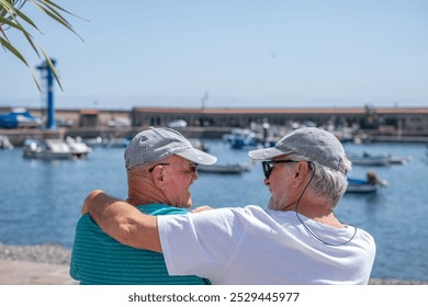 Rear view of cheerful senior couple of men friends sitting on a bench face the sea port laughing and looking at each other, two elderly pensioner males enjoying healthy lifestyle in outdoors - Powered by Shutterstock