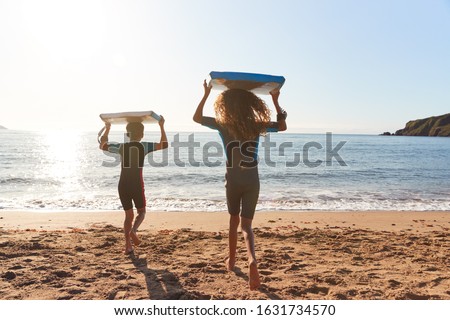 Rear View Of Children In Wetsuits Carrying Bodyboards On Summer Beach Vacation Having Fun By Sea